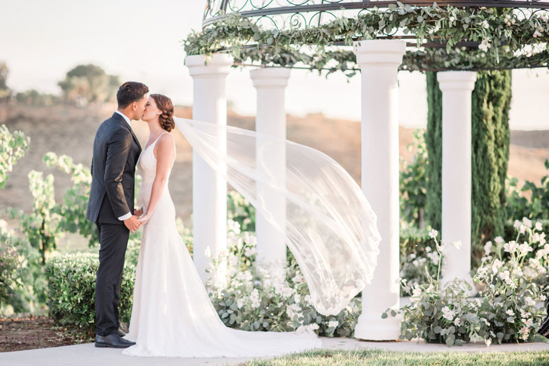 Bride and groom kissing next to their wedding gazebo. Bride's veil is blowing in the wind.